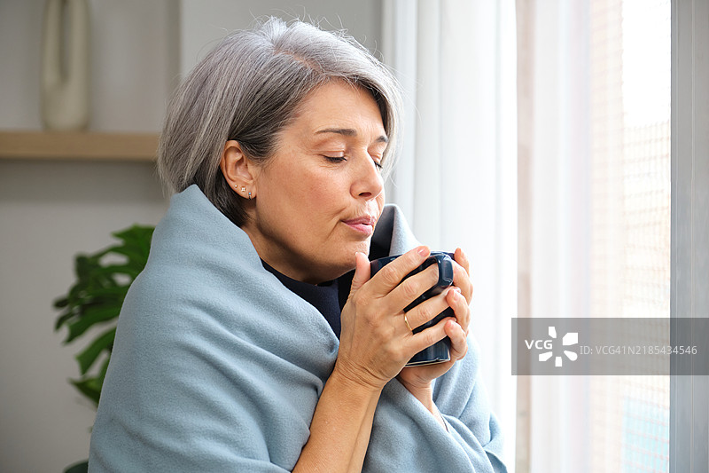 裹着毛毯的成熟的女性在窗边享受热饮图片素材