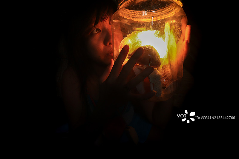 亚洲女孩在黑暗中握着一个发光的罐子，营造出神秘而沉思的氛围图片素材