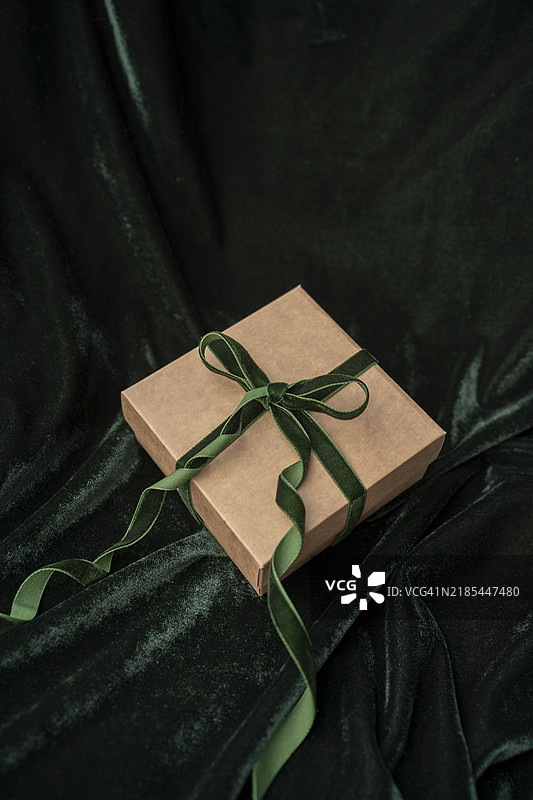 一个圣诞礼物盒，上面系着绿色天鹅绒丝带和蝴蝶结，背景是深绿色天鹅绒。图片素材