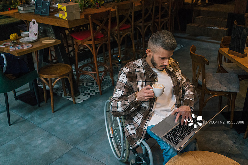 一位年轻男子坐在咖啡馆里，使用轮椅，悠闲地喝着咖啡，认真地在笔记本电脑上写邮件，享受着周围的氛围。一位残疾人士在咖啡馆里，愉快地享受着他的咖啡，心情愉悦。图片素材