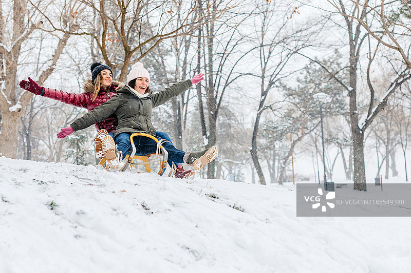 两个女性朋友在冬季的雪林中坐着雪橇下坡图片素材