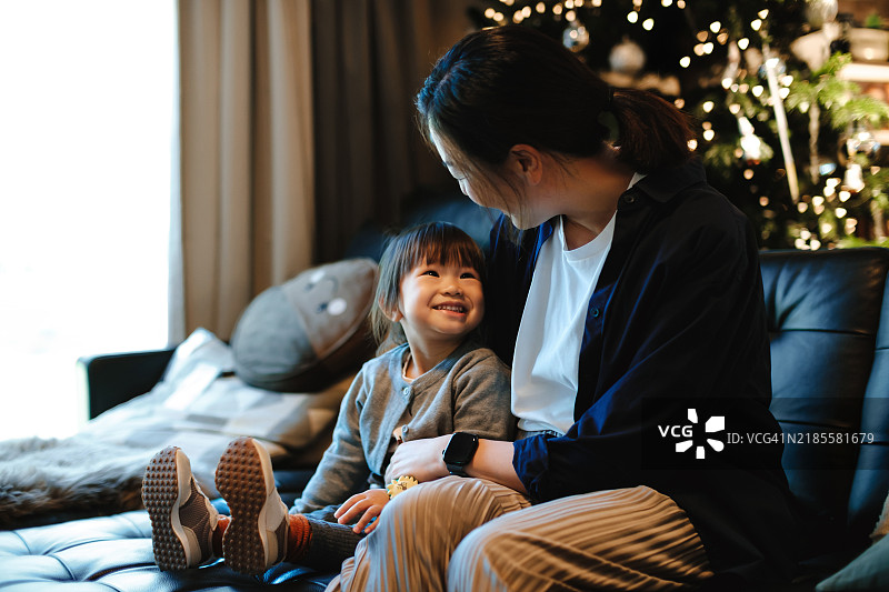 年轻的亚洲母亲与可爱的幼女在温馨的家中度过圣诞季，坐在客厅的沙发上，放松地拥抱着女孩，旁边是一棵装饰华丽的圣诞树。家庭的爱、和谐与团聚。图片素材