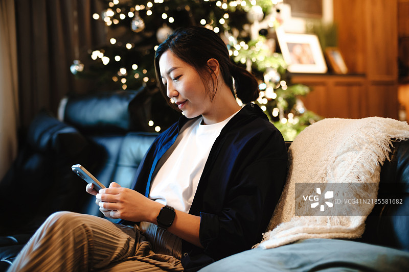 微笑的年轻亚洲女性坐在客厅的沙发上，享受在线购物，使用智能手机，旁边是一棵装饰华丽的圣诞树。圣诞购物、购买礼物、为圣诞节做准备。圣诞节和新年庆祝活动。图片素材