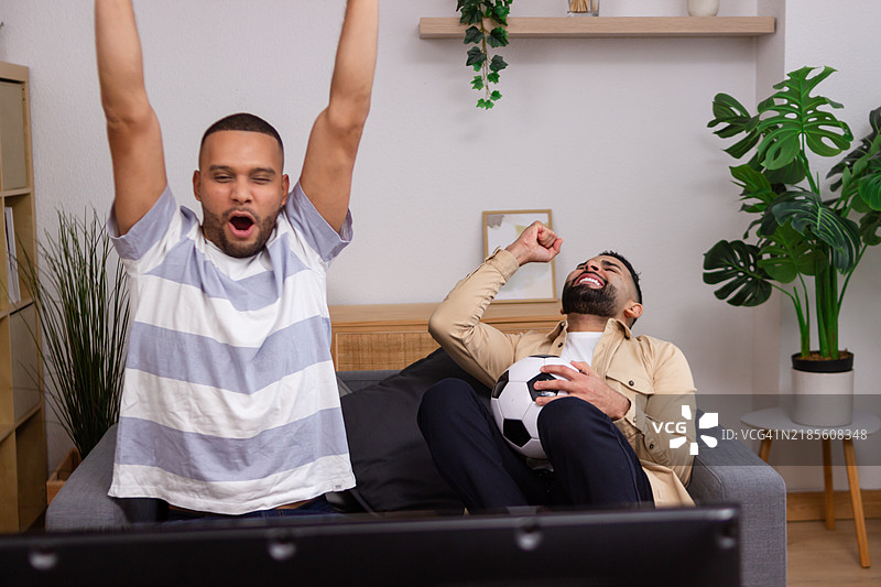 两个男性朋友在家看足球比赛，庆祝进球。图片素材