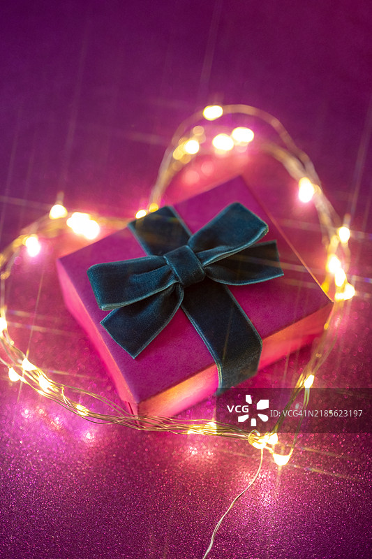 一个精美包装的蓝色礼品盒，系着红色丝带，位于心形发光灯饰的中央，非常适合情人节或生日等庆祝活动。图片素材