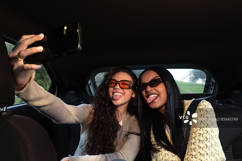 两位女性朋友在车里摆姿势自拍，调皮地吐舌头并戴着太阳镜。图片素材