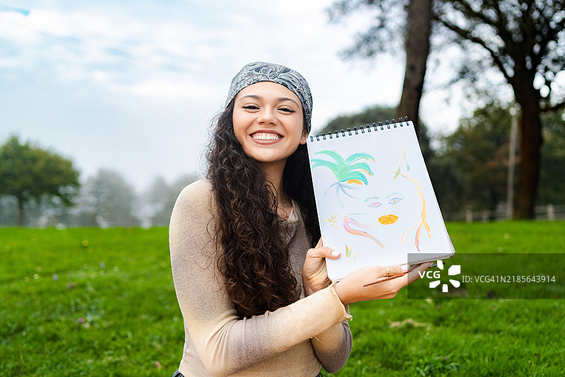 年轻女性在公园里微笑着展示她的彩色画作，享受着大自然中的爱好图片素材