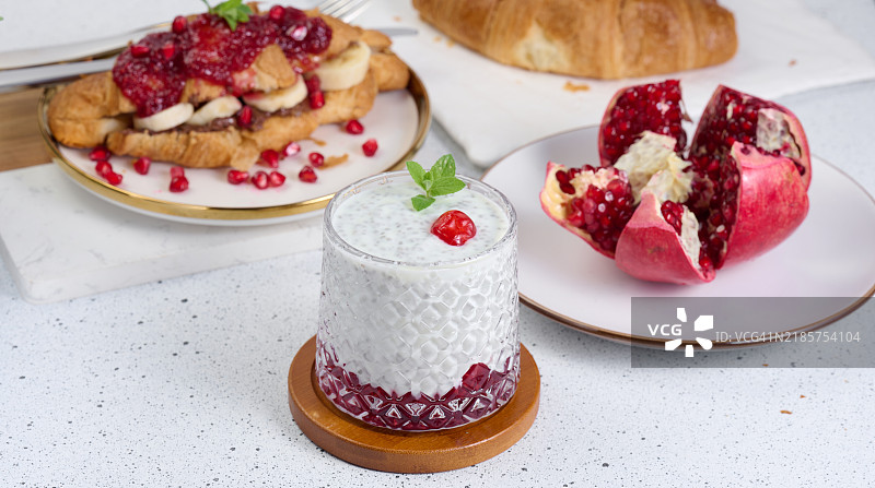 装有奇亚籽和酸奶甜点的玻璃杯，上面装饰着绿色薄荷叶，放在白色桌子上。图片素材