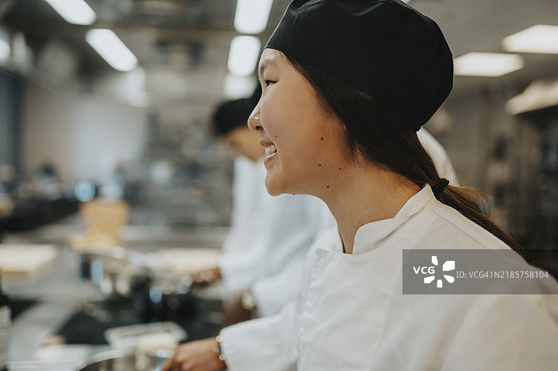 侧面视图的微笑少女学员，戴着厨师帽，在烹饪学校学习图片素材