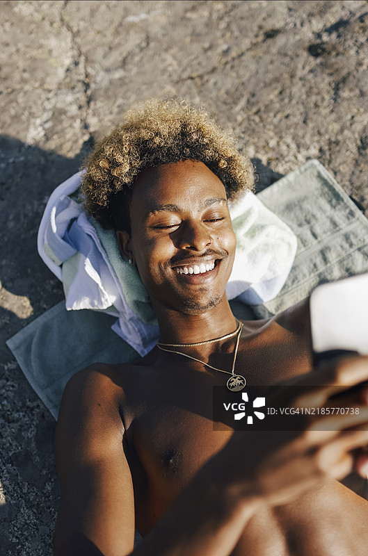 高角度视图，微笑的年轻男子赤裸上身，躺在岩石上使用智能手机。图片素材