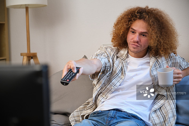 年轻男子正在看电视，手里拿着遥控器和一个杯子。图片素材