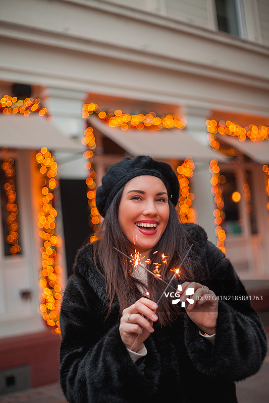 快乐的年轻黑发女性穿着仿毛皮外套和贝雷帽，在欧洲城市中心用烟花庆祝新年。冬季假期。新年。图片素材