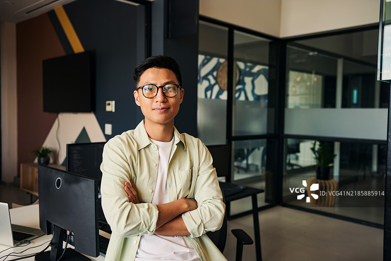 年轻的亚洲计算机程序员坐在现代办公室里，双臂交叉。图片素材