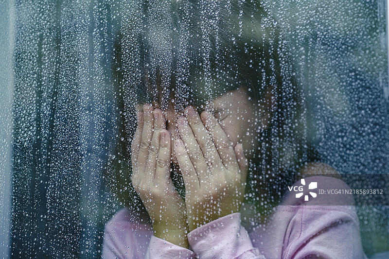 伤心的孩子在雨窗后哭泣，用手遮住脸。图片素材