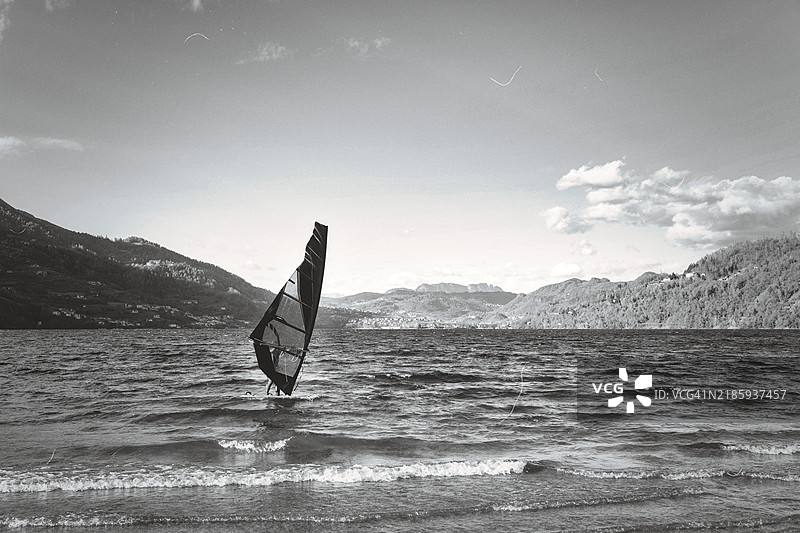 黑白照片，展示了一名风帆冲浪者在意大利特伦蒂诺-阿尔托-阿迪杰的卡尔多纳佐湖上冲浪，拍摄于2024年11月的一个风大的日子。图片素材
