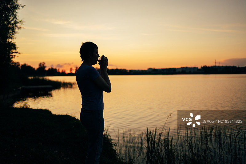 湖边日落时分，一位女性的剪影在享受饮品，感受大自然的宁静。图片素材