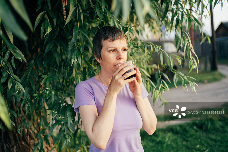 在日落时分，宁静的女人在柳树下喝着杯子里的饮品图片素材