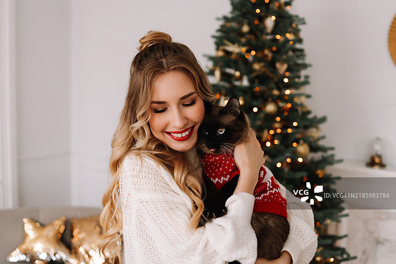 一位美丽的女性在冬季十二月的室内，拥抱着她的宠物猫，背景是装饰华丽的圣诞树。微笑的女孩与她的家养动物的肖像。图片素材