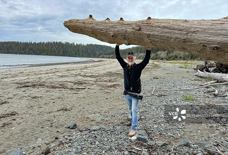 一位老年男子站在海滩上，身旁有一根漂流木，温哥华岛，加拿大不列颠哥伦比亚省温哥华。图片素材