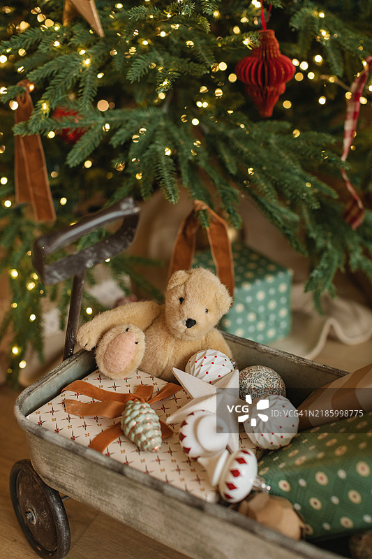 特写玩具小车，装满了包裹好的圣诞礼物、泰迪熊和各种装饰品，旁边是一棵闪亮的圣诞树。图片素材
