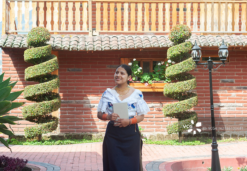 年轻的拉丁美洲房地产顾问微笑着，手持平板电脑，站在一栋有螺旋修剪树的房子外。图片素材