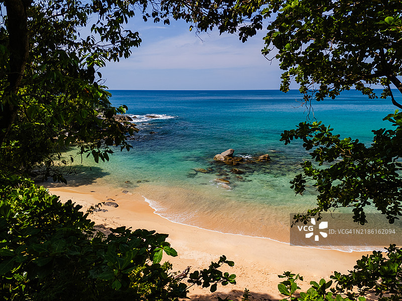 热带海滩的景色，金色沙滩和碧绿色海水，周围环绕着郁郁葱葱的绿叶和零星的岩石。图片素材