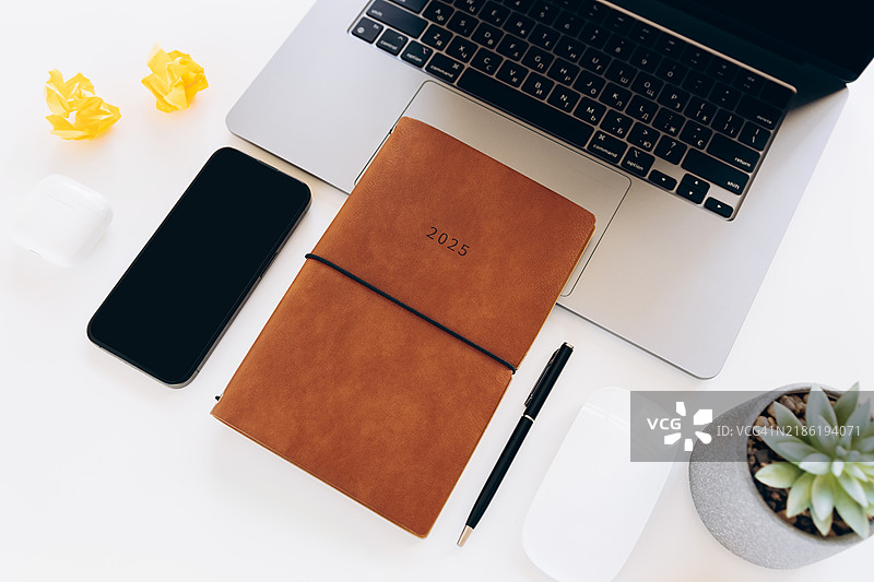现代工作空间平铺，包含2025年计划本、笔记本电脑、智能手机、无线鼠标、钢笔、多肉植物和皱巴巴的纸张，摆放在干净的白色桌面上。极简主义办公设置，旨在提高生产力和规划。图片素材