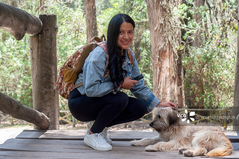 年轻女性背着背包，蹲下身来抚摸一只毛茸茸的狗，享受着在宁静森林中的独自旅行冒险。图片素材