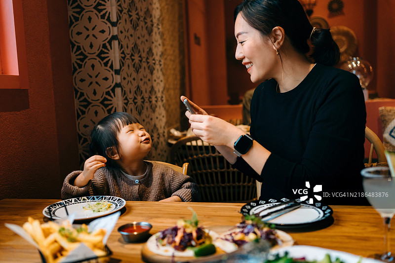 年轻的亚洲母亲在时尚的墨西哥餐厅里用智能手机拍摄她可爱的小女儿，同时享受美食。家庭外出就餐。母女亲密时刻。捕捉回忆。家庭、爱与团聚的概念。图片素材