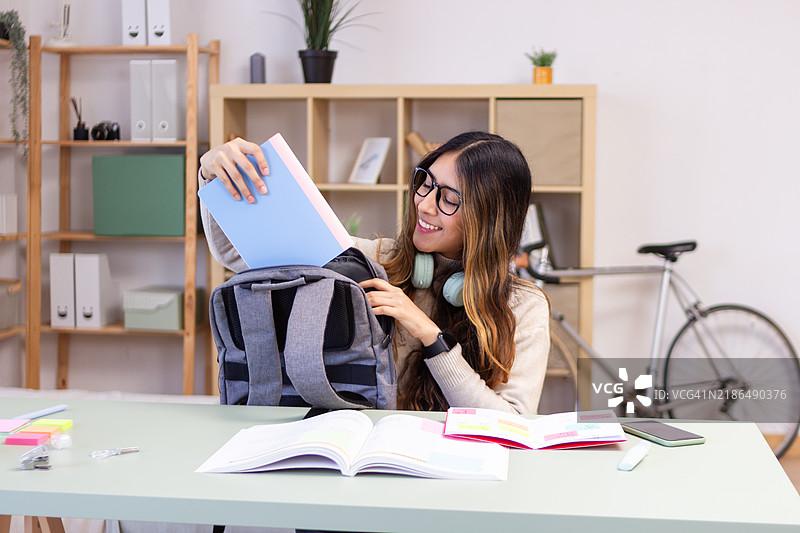 年轻的拉丁裔女性在家办公桌上把书放进背包里图片素材