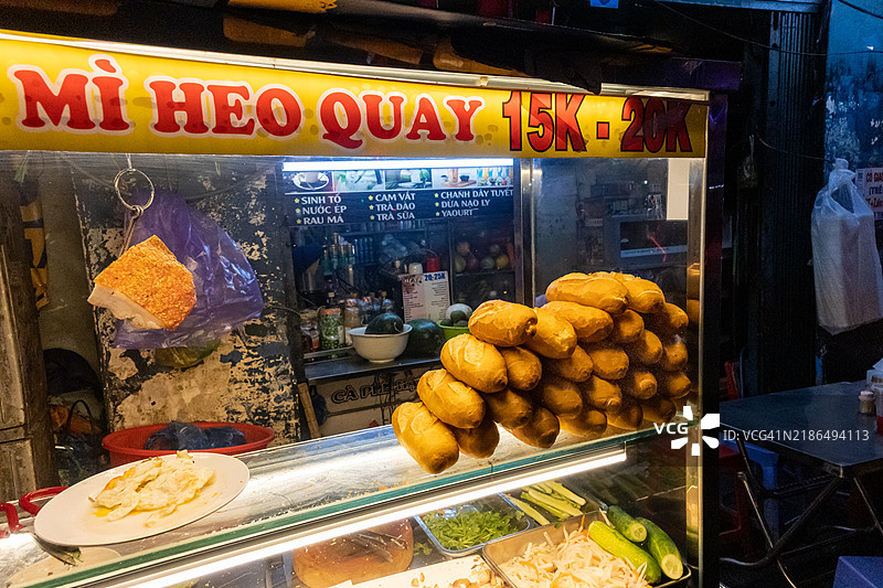 夜晚的胡志明市，一个街头摊位展示着越南烧肉法棍（Banh mi heo quay），摊位上堆放着法棍面包。图片素材