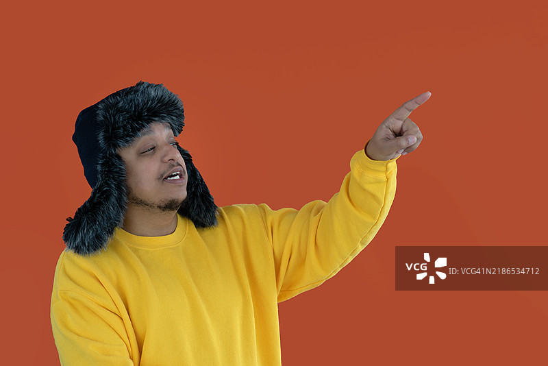 一个戴着乌莎卡帽的人站在橙色的纯色背景上，用食指指向上方。图片素材