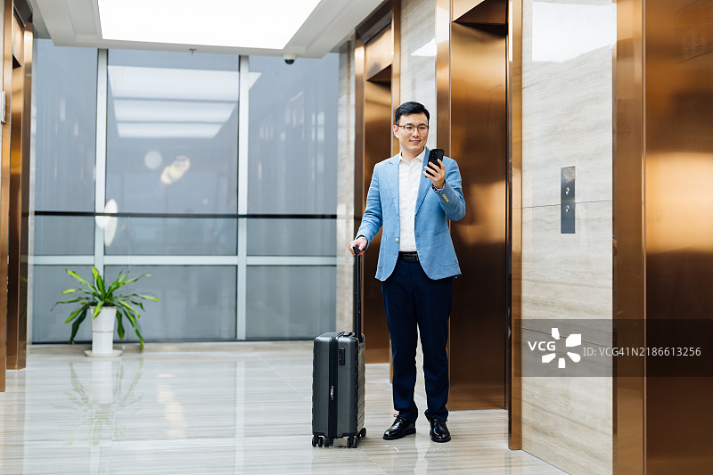 亚洲商人使用智能手机，拖着行李箱靠近电梯。图片素材