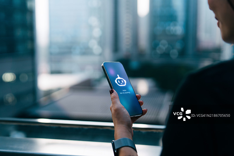 年轻的亚洲女商人在城市露台上站立，背对现代摩天大楼，正在与智能手机上的虚拟助手ChatGPT AI聊天机器人应用交谈。虚拟现实和增强现实技术的概念。人们、人工智能、创新和科技。图片素材