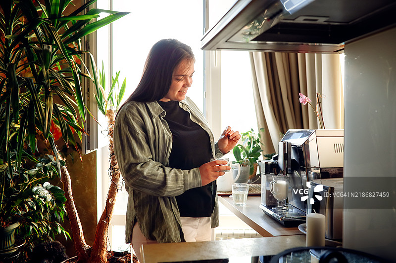 一位丰满女性在昏暗温馨的厨房中享受宁静的早晨仪式，使用咖啡机制作咖啡。图片素材