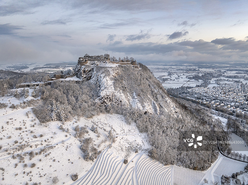 雪覆盖的赫高火山霍亨特维尔的鸟瞰图,冬季寒冷的早晨,日出前景,周围是结霜的树木,冬季风景中,右侧是巴登-符腾堡州康斯坦茨地区的辛根镇,德国最大的城堡遗址。图片素材