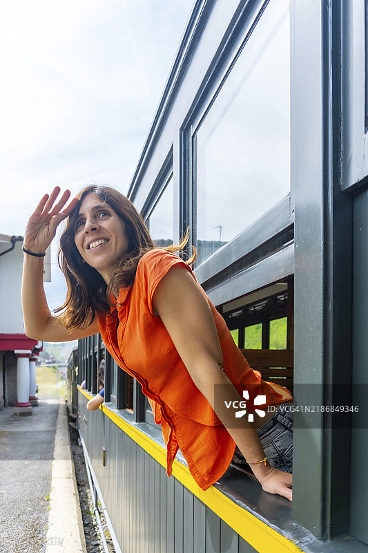 一位穿着橙色衬衫的女性正在火车窗外望去。她面带微笑，向某人挥手。图片素材