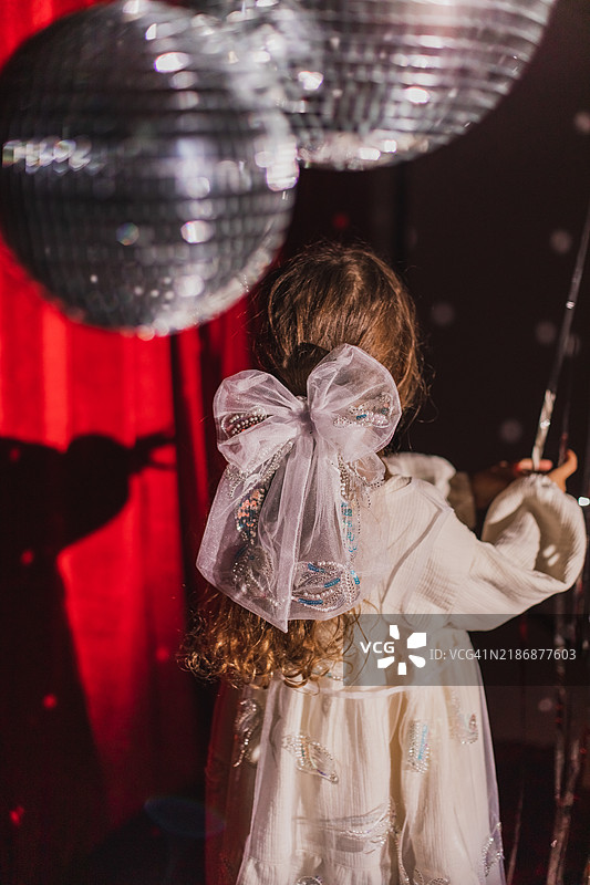 美丽的小女孩穿着亮片裙，站在迪斯科球和银色闪光带之间，背景是红色天鹅绒。图片素材