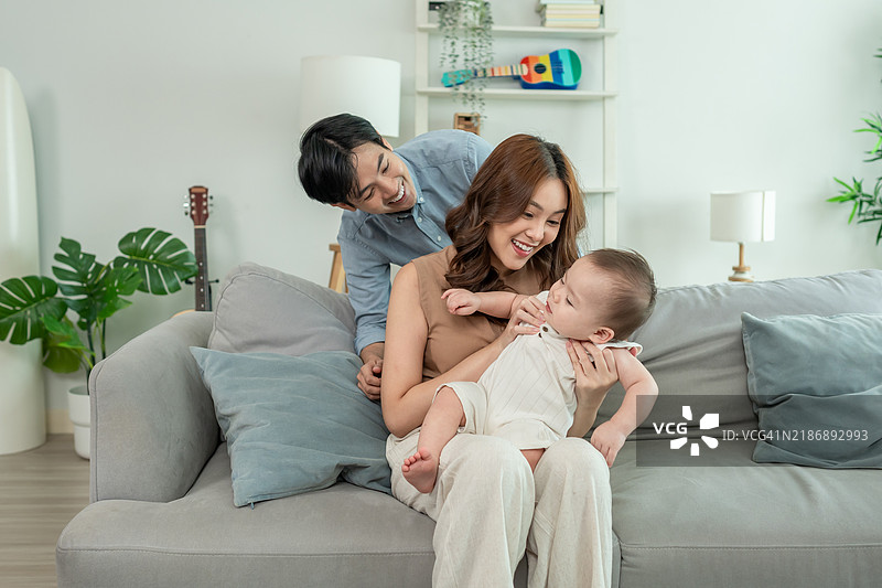 亚洲有吸引力的父母温柔地抱着他们快乐的男婴。可爱的小男孩在家里的沙发上与爸爸妈妈愉快互动，享受着玩耍的时光。图片素材