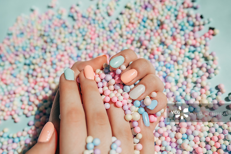 女性的手指上涂有时尚多彩的法式美甲，放在一个由浅绿色和浅粉色部分组成的柔和背景上，周围散落着许多圆形纸质彩屑。设计留白。平铺风格。图片素材