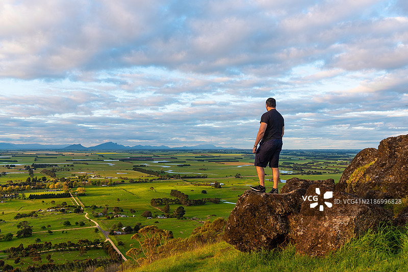 一个男人站在罗斯山顶的背影，俯瞰着农田，头顶上有美丽的云彩。图片素材