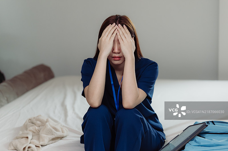 压力大的亚洲护士坐在卧室的床上，双手捂住脸。图片素材