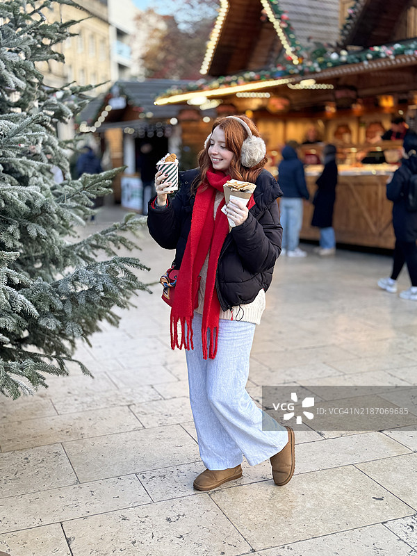 穿着黑色羽绒服、红色围巾和仿毛耳罩的迷人红发女性，手里拿着一次性纸杯热巧克力和纸筒装的法式可丽饼，背景是圣诞树和户外圣诞市场。图片素材