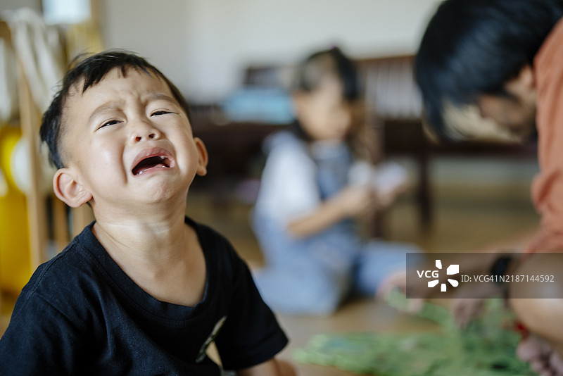 一个哭泣的亚洲幼儿在镜头前，背景中家人们正在玩耍，捕捉到家庭环境中的真实育儿时刻。图片素材