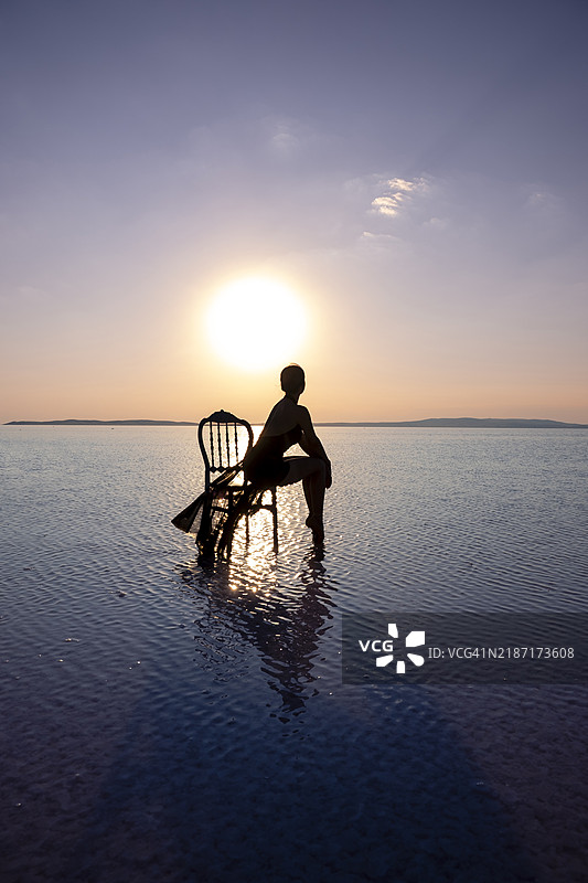 女人坐在浅水中的椅子上，夕阳西下。图片素材