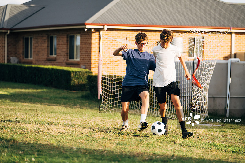 两个少年男孩在郁郁葱葱的草地或后院踢足球，积极参与一场竞争激烈的比赛，场景设在住宅后院。图片素材