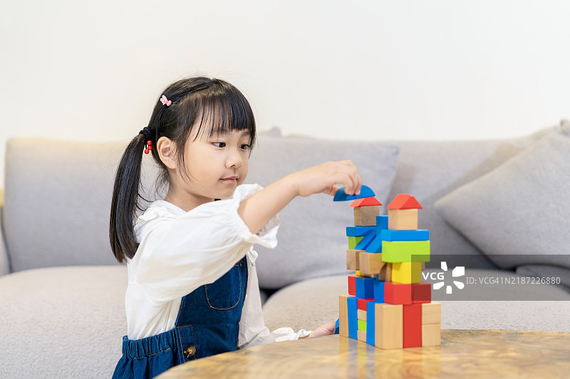 一个可爱的亚洲小女孩蹲在桌子旁，正在家里玩木制积木。图片素材