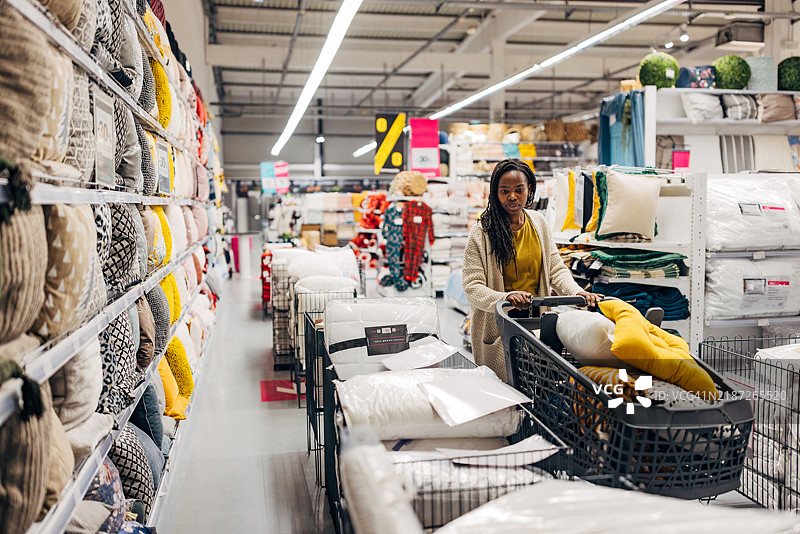 一位黑人女性在百货商店寻找高质量、舒适且柔软的枕头。她推着购物车在商店里挑选要购买的枕头。图片素材