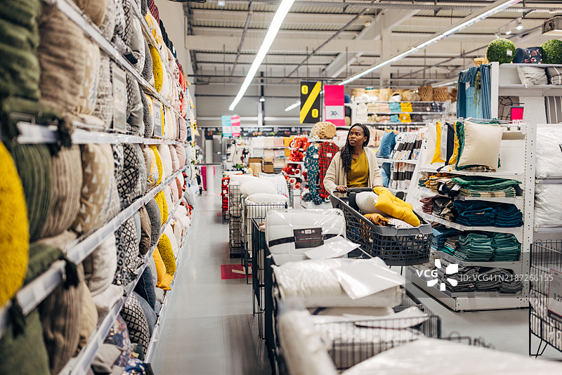 一位黑人女性推着购物车穿过百货商店的一部分，那里出售各种材质、颜色和图案的枕头。她在决定购买哪些枕头。图片素材