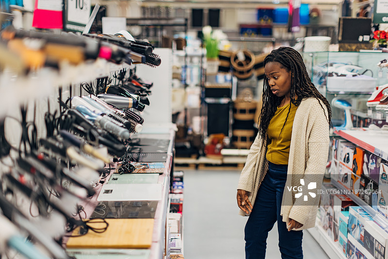 一位无忧无虑的黑人女性穿着休闲服装，在百货商店购买吹风机。她站在一个货架前，货架上展示着不同类型和品牌的吹风机和电动刷子。图片素材